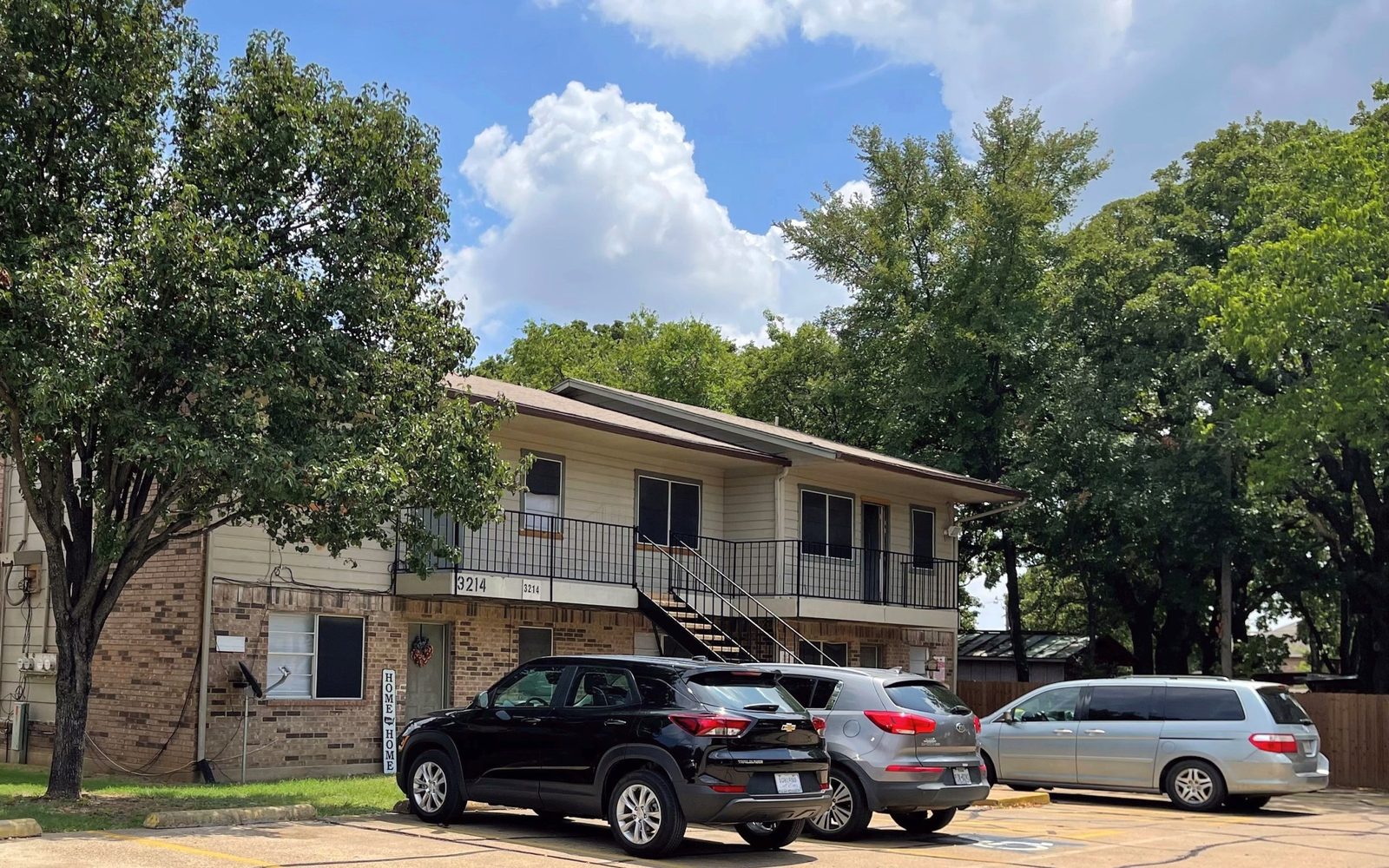 Two-story brick apartment building under large oak trees with cars parked in front