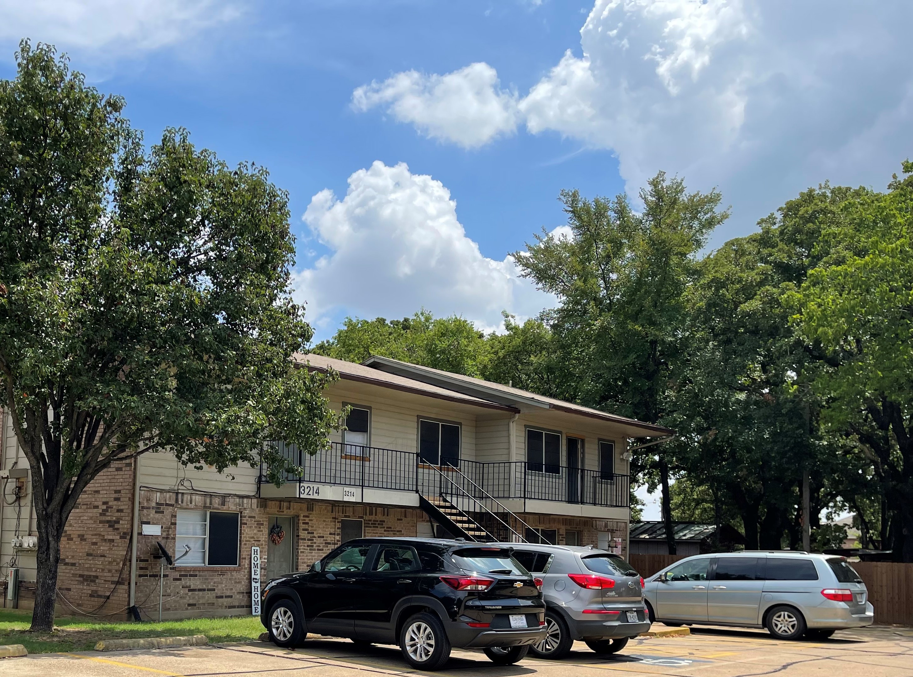 Two-story apartment building under large oak trees with cars parked in front